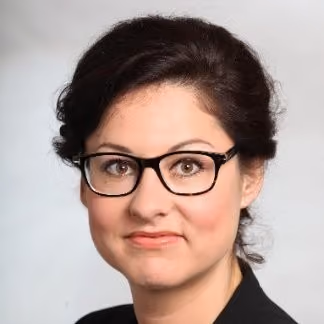 Frau mit dunkelbraunen Haaren und schwarzen Brillengestell vor hellgrauem Hintergrund.