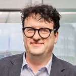 Porträt eines Mannes mit dunklen lockigen Haaren und Brille vor einem Fenster.
