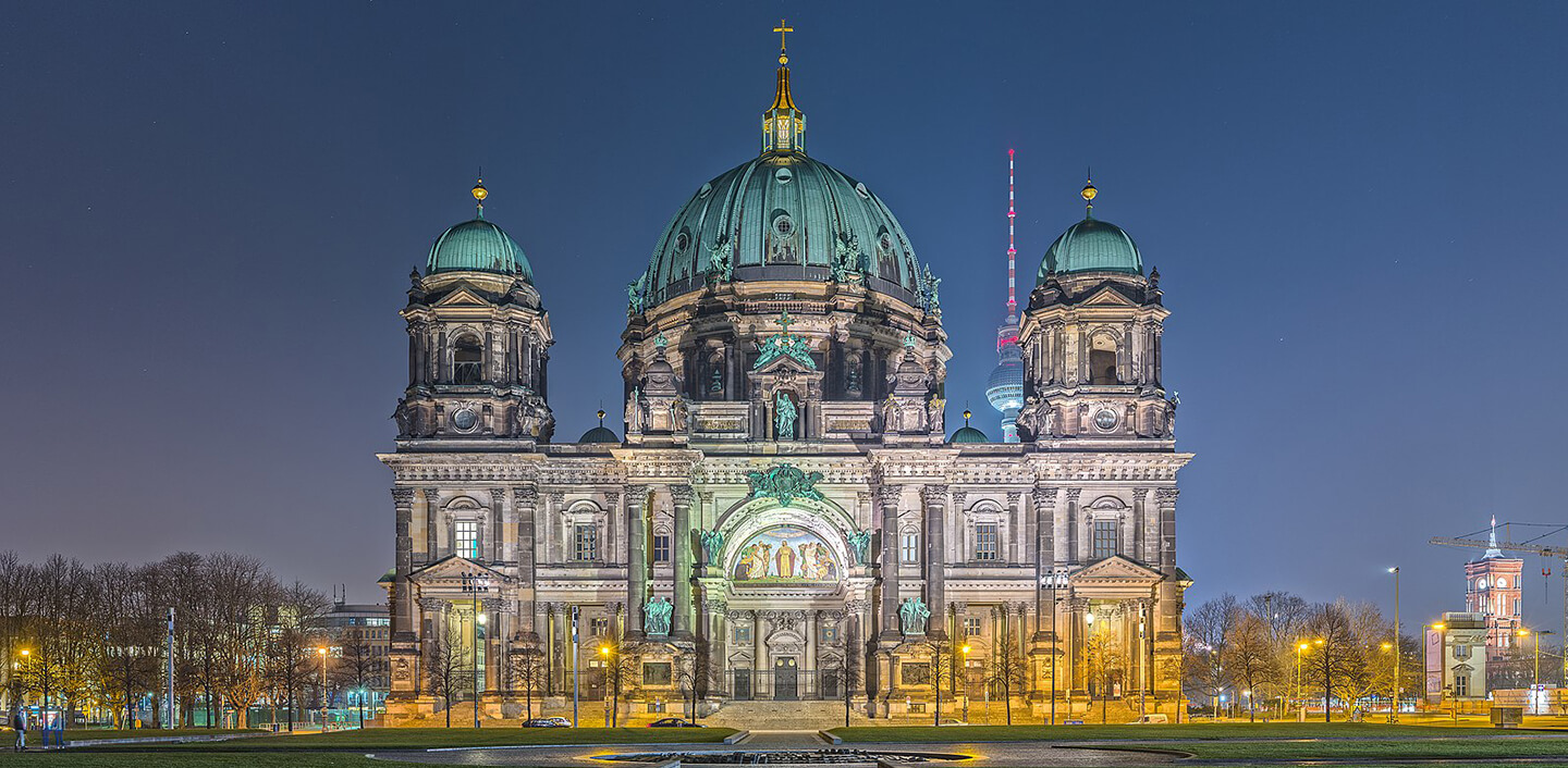 Berliner Dom im hellen Licht bei Nacht mit Fernsehturm im Hintergrund und Bäumen rechts und links.