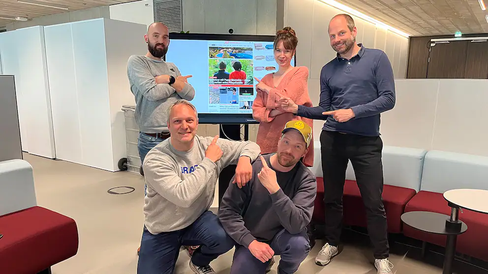 Fünf Personen zeigen zusammen auf einen Bildschirm mit einer Webseite in einem modernen Büro.