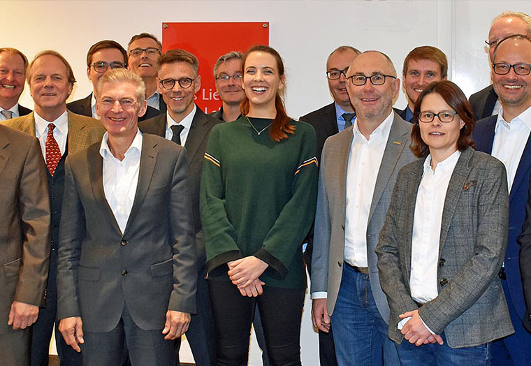 Gruppe von Menschen in Anzügen und Freizeitkleidung, die vor weißer Wand mit rotem Schild stehen.
