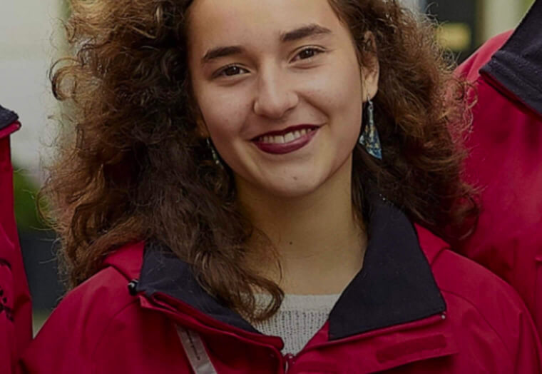 Junge Frau mit lockigem Haar, roter Jacke und dunklem Lippenstift lächelt in die Kamera.