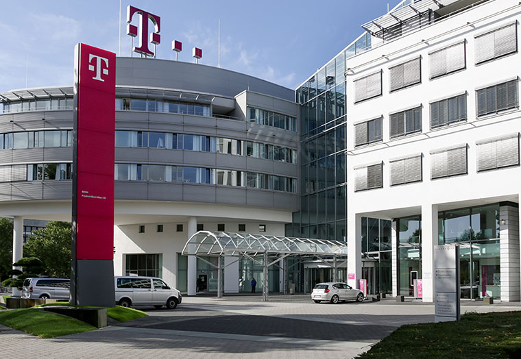 Gebäude der Telekom mit großem Logo, Einfahrt und mehreren parkenden Autos bei klarem Himmel.