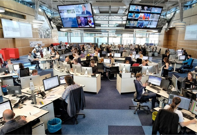 Großes Nachrichtenbüro mit vielen Mitarbeitern an Computern und zwei Bildschirmen an der Decke.