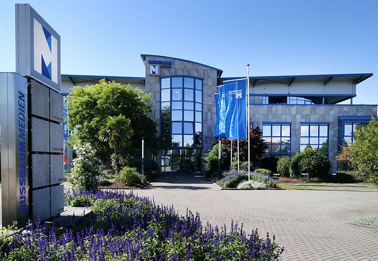 Bürogebäude von Nusbaum Medien mit Glasfenstern, blauen Fahnen und blühenden Pflanzen im Vordergrund.