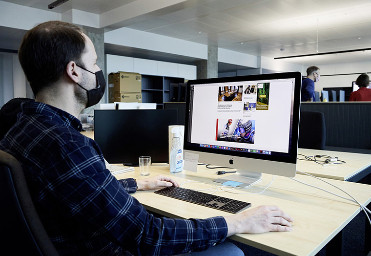 Mann mit Maske arbeitet an einem Schreibtisch mit iMac in einem offenen Büro.