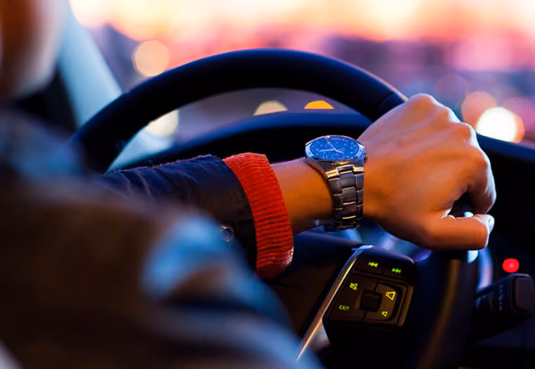 Hand with wristwatch on steering wheel inside car at dusk with blurred lights outside.
