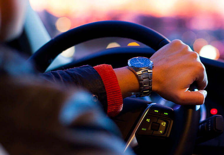 Hand with wristwatch on steering wheel inside car at dusk with blurred lights outside.