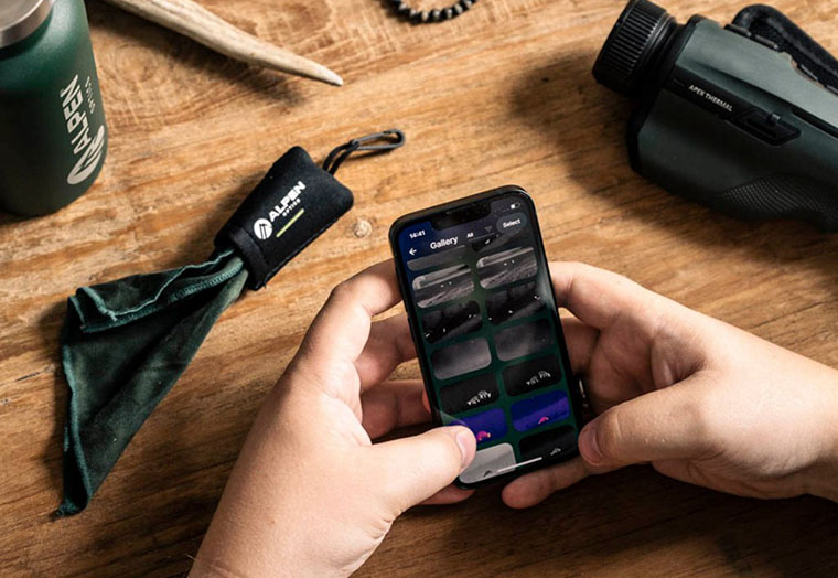 Person hält Smartphone mit geöffnetem Foto-Viewer auf hölzernem Tisch neben Alpen Optics Brillenbeutel und Gerät.