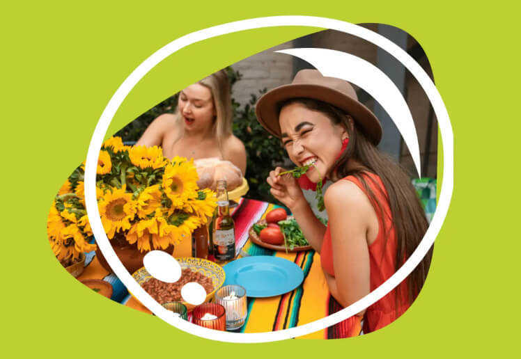Zwei Frauen lachen und essen Salat bei einem sonnigen Tisch mit Sonnenblumen.