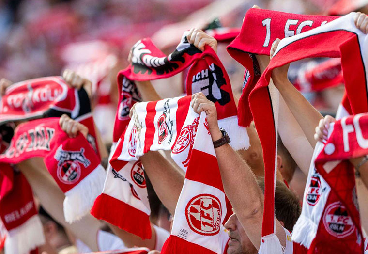 Fans halten rote und weiße 1. FC Köln Schals bei einem Fußballspiel hoch.