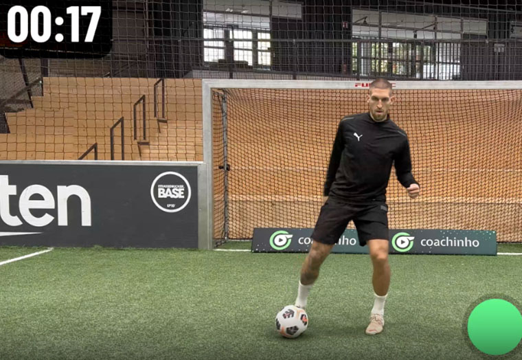 Ein Fußballspieler in schwarzem Trikot dribbelt vor einem Tor auf Kunstrasen mit Timer 00:17.