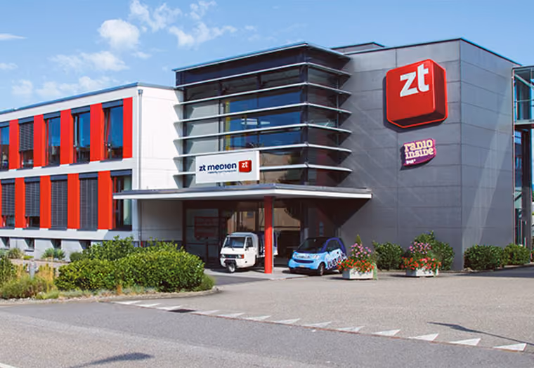 Modernes Bürogebäude mit roten Akzenten und zwei Autos vor dem Eingangsbereich an einem sonnigen Tag.