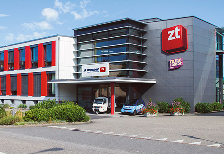 Modernes Bürogebäude mit roten Akzenten und zwei Autos vor dem Eingangsbereich an einem sonnigen Tag.