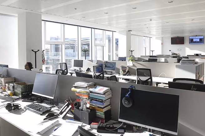 Helles Büro mit Reihen von Schreibtischen, Computern, Bücherstapeln und mehreren Stühlen.