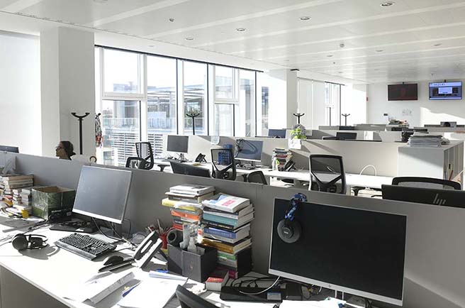 Helles Büro mit Reihen von Schreibtischen, Computern, Bücherstapeln und mehreren Stühlen.