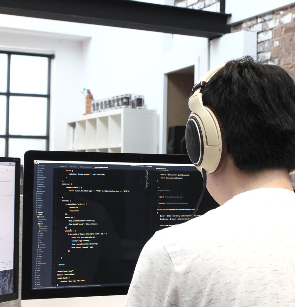 Person mit Kopfhörern arbeitet an Computer mit Programmiercode auf Bildschirm in Büro.