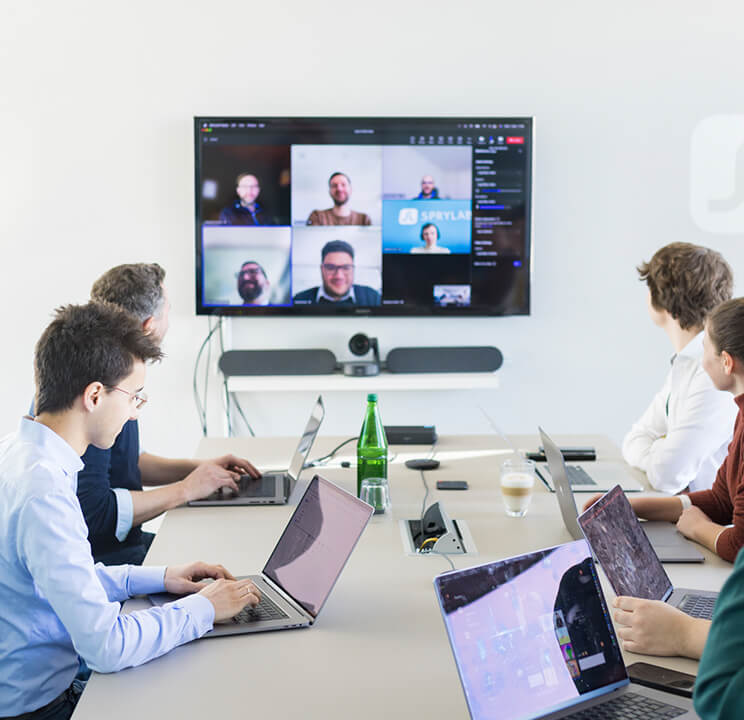 Mehrere Personen sitzen an einem Konferenztisch mit Laptops und nehmen an einer Videoanrufbesprechung teil.