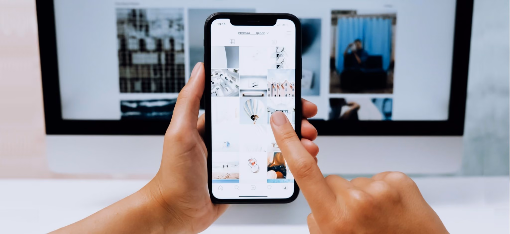 Person nutzt eine Social-Media-App auf dem Smartphone vor einem Computerbildschirm.