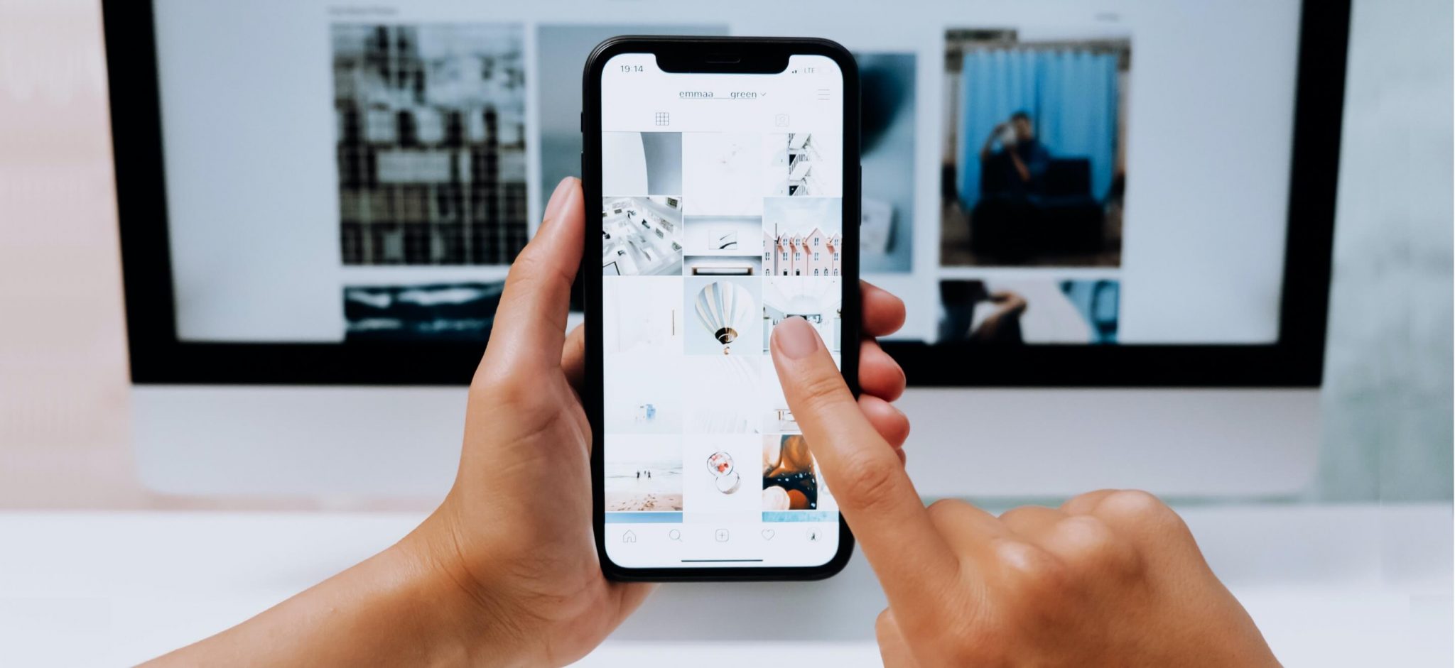 Person nutzt eine Social-Media-App auf dem Smartphone vor einem Computerbildschirm.