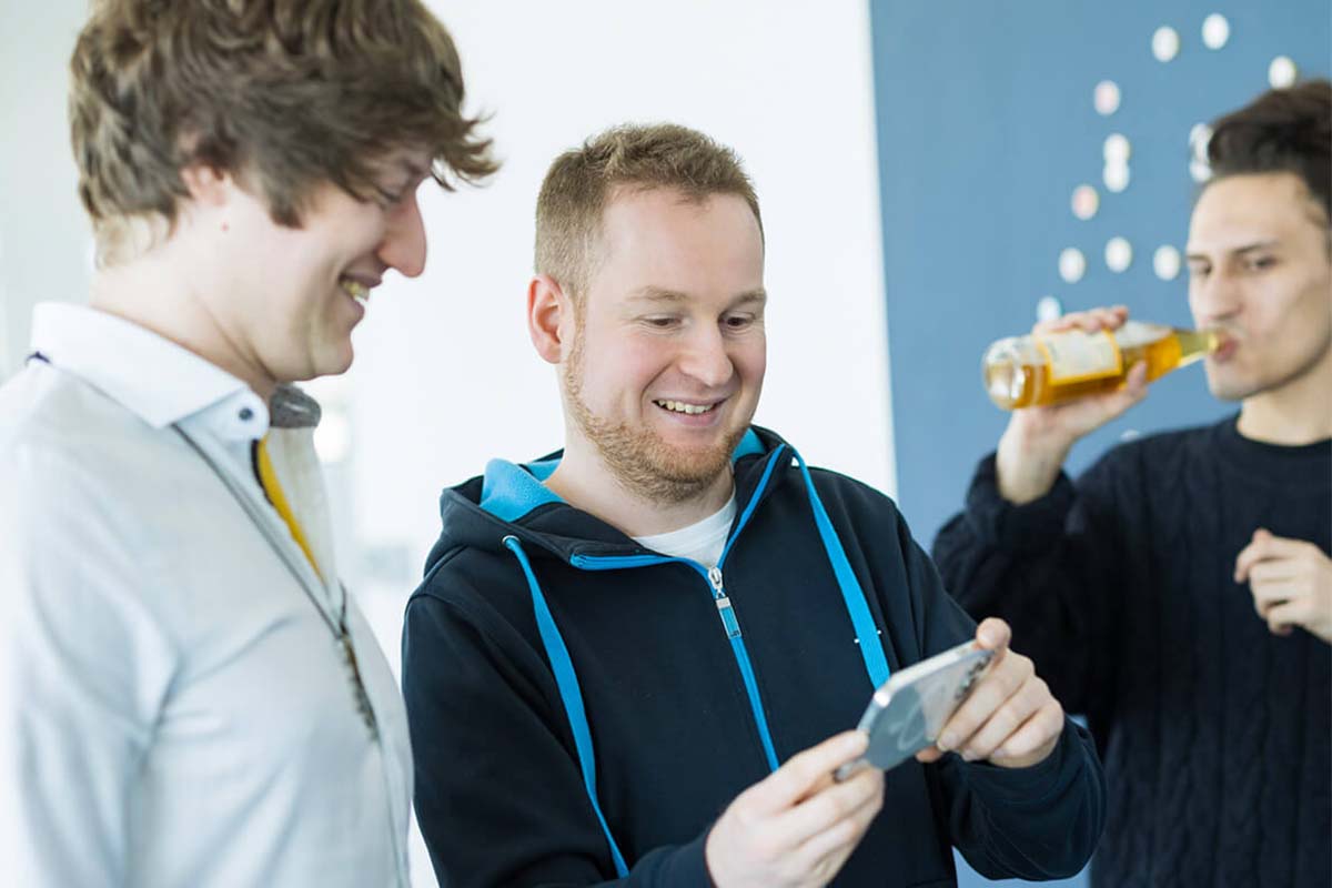 Drei Männer, zwei lachen und schauen auf ein Smartphone, der dritte trinkt ein Bier.