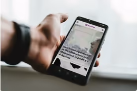 Hand hält Smartphone, darauf Webseite mit textlastigem Artikel und einem Foto zu sehen.