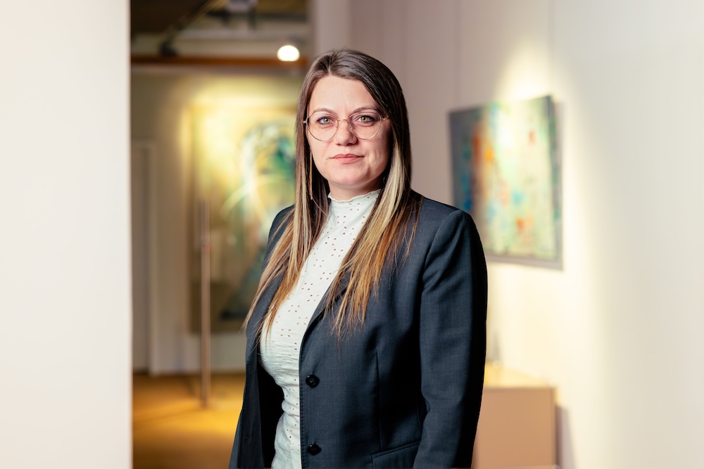 Frau mit Brille und langem Haar trägt schwarzen Blazer vor Kunstwerken in einem Galerieflur.