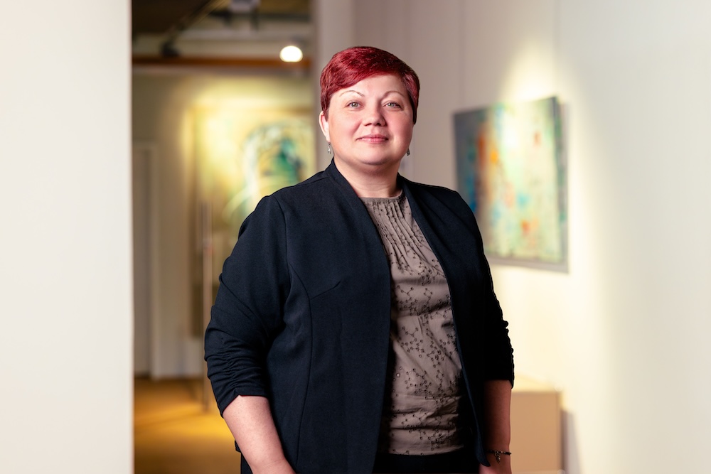 Frau mit kurzen roten Haaren trägt schwarze Jacke und graues Oberteil steht in heller Galerie.
