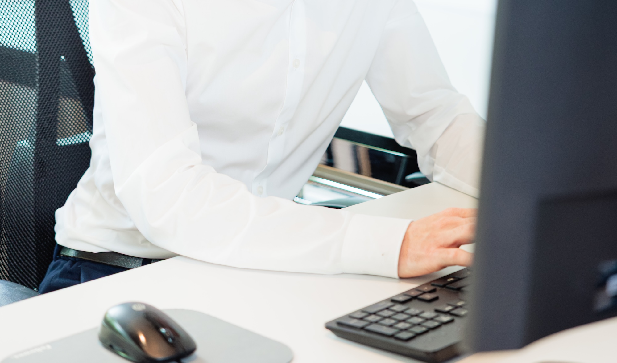 Eine Person in weißem Hemd arbeitet an einem Computer mit Tastatur und Maus auf weißem Schreibtisch.