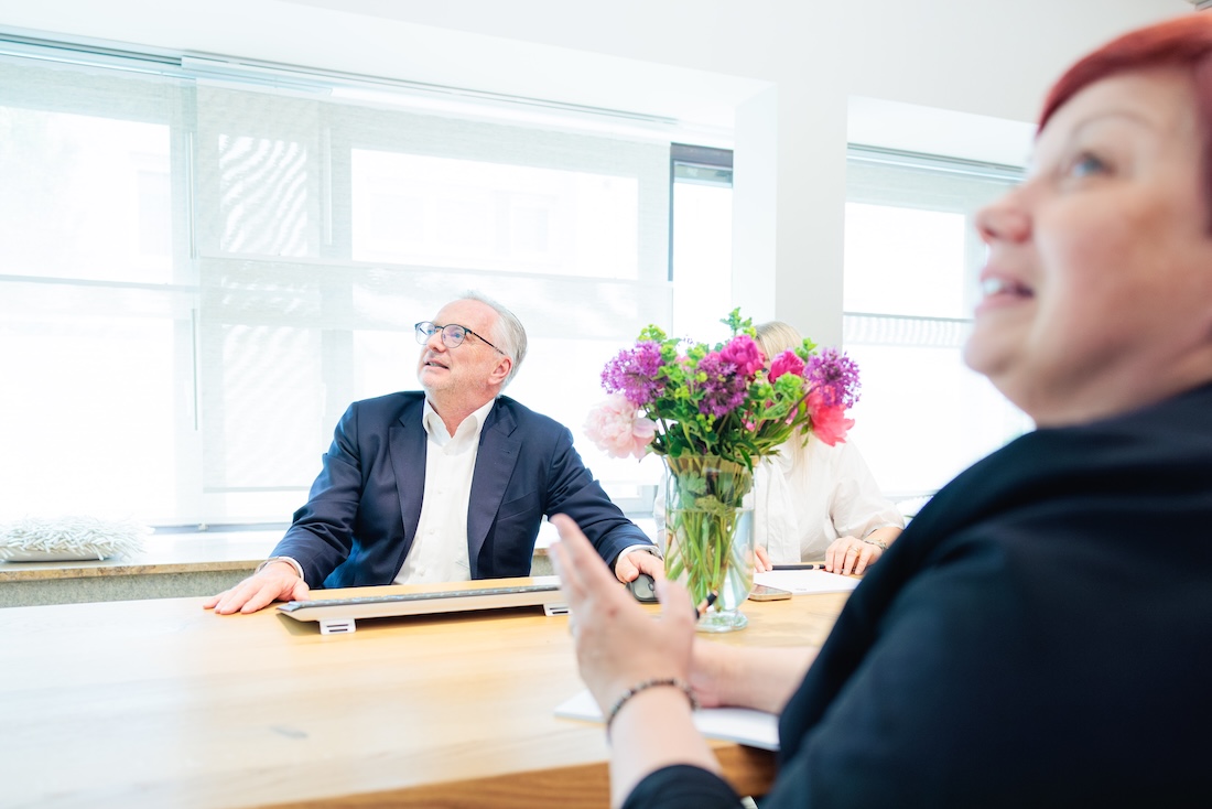 Ein älterer Mann in Anzug und eine Frau im Büro führen ein Gespräch am Holztisch mit Blumen in der Mitte.