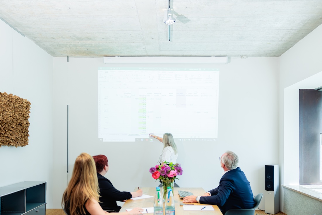 Frau präsentiert Daten an Wand, drei Personen sitzen an Tisch und schauen zu im modernen Büro.