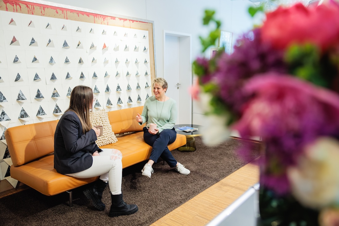 Zwei Frauen sitzen auf einer orangefarbenen Couch und trinken Kaffee im modernen Büro.