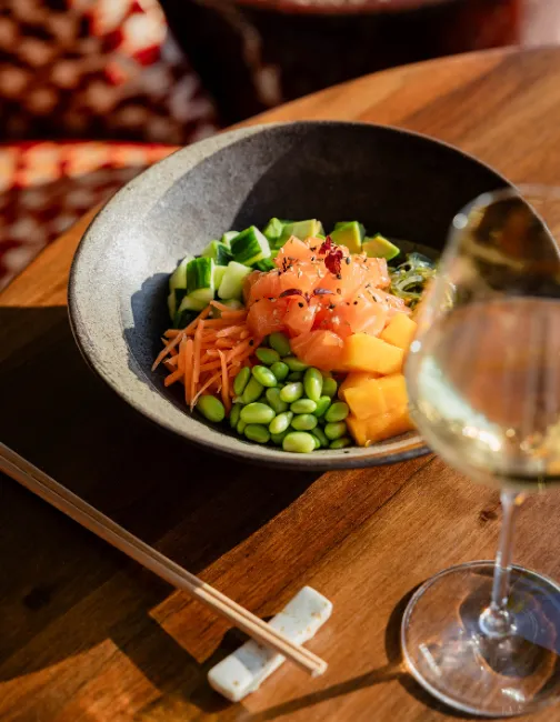 Salmon poke bowl with cucumber, carrot, edamame, and mango, served on a wooden table with a glass of white wine.