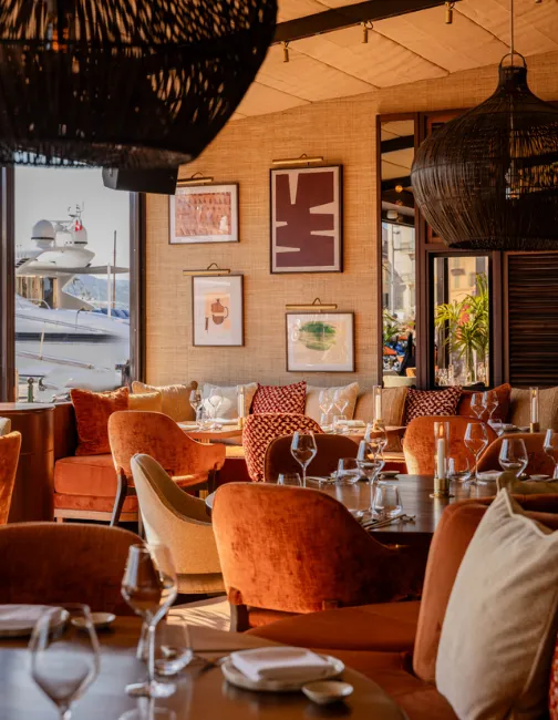 Light-filled dining room featuring orange velvet armchairs around set tables, black rattan pendant lights, and a view of the harbor with a yacht.