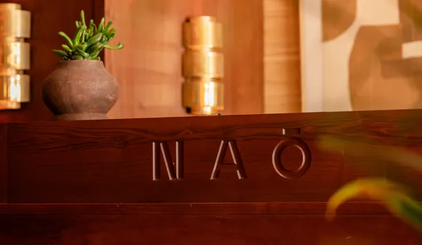 Logo NAO gravé sur un comptoir en bois, avec applique dorée et plante succulente en arrière-plan.