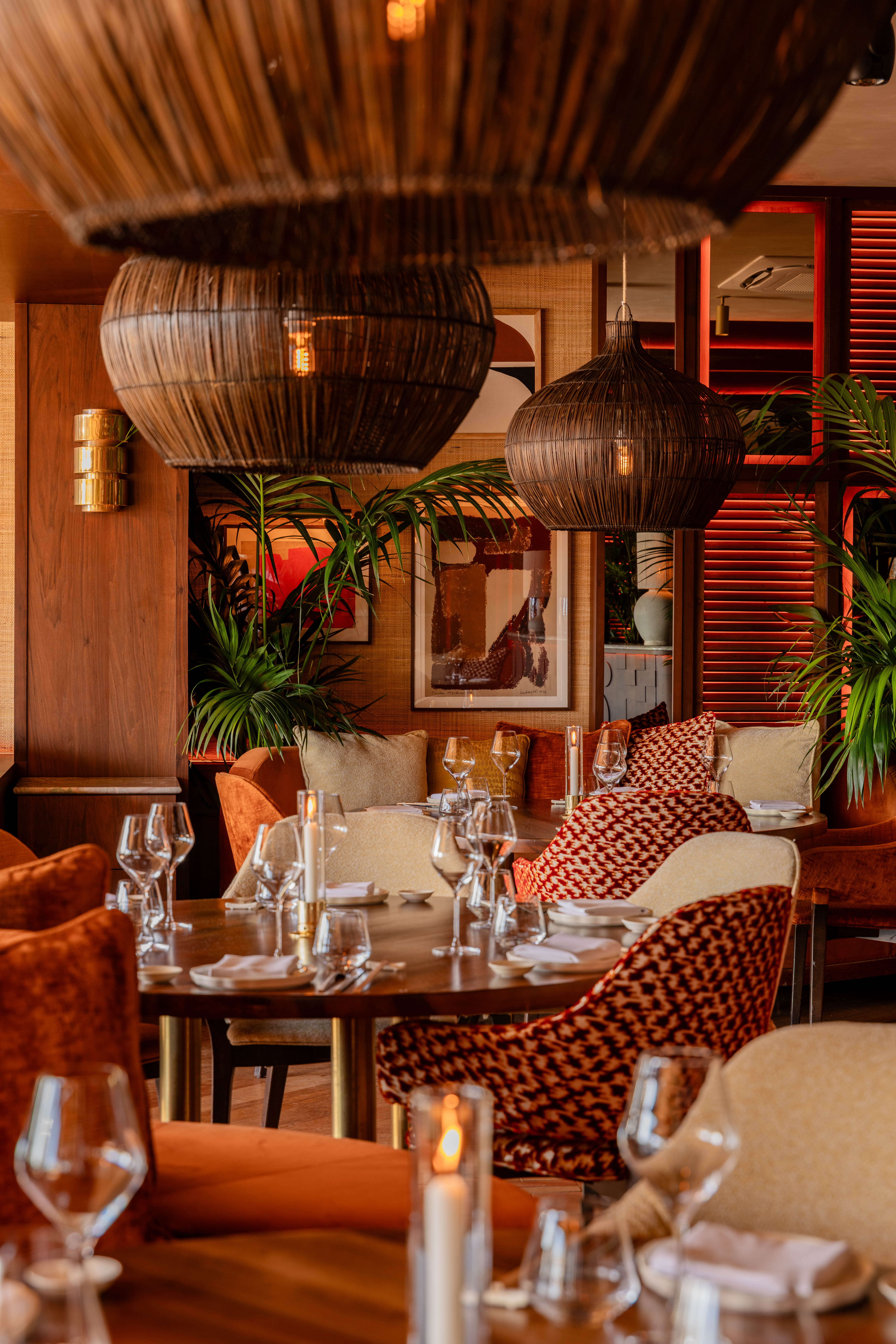Salle de restaurant chaleureuse avec suspensions en rotin tressé, tables rondes dressées, fauteuils en velours orange et persiennes rétroéclairées en rouge.