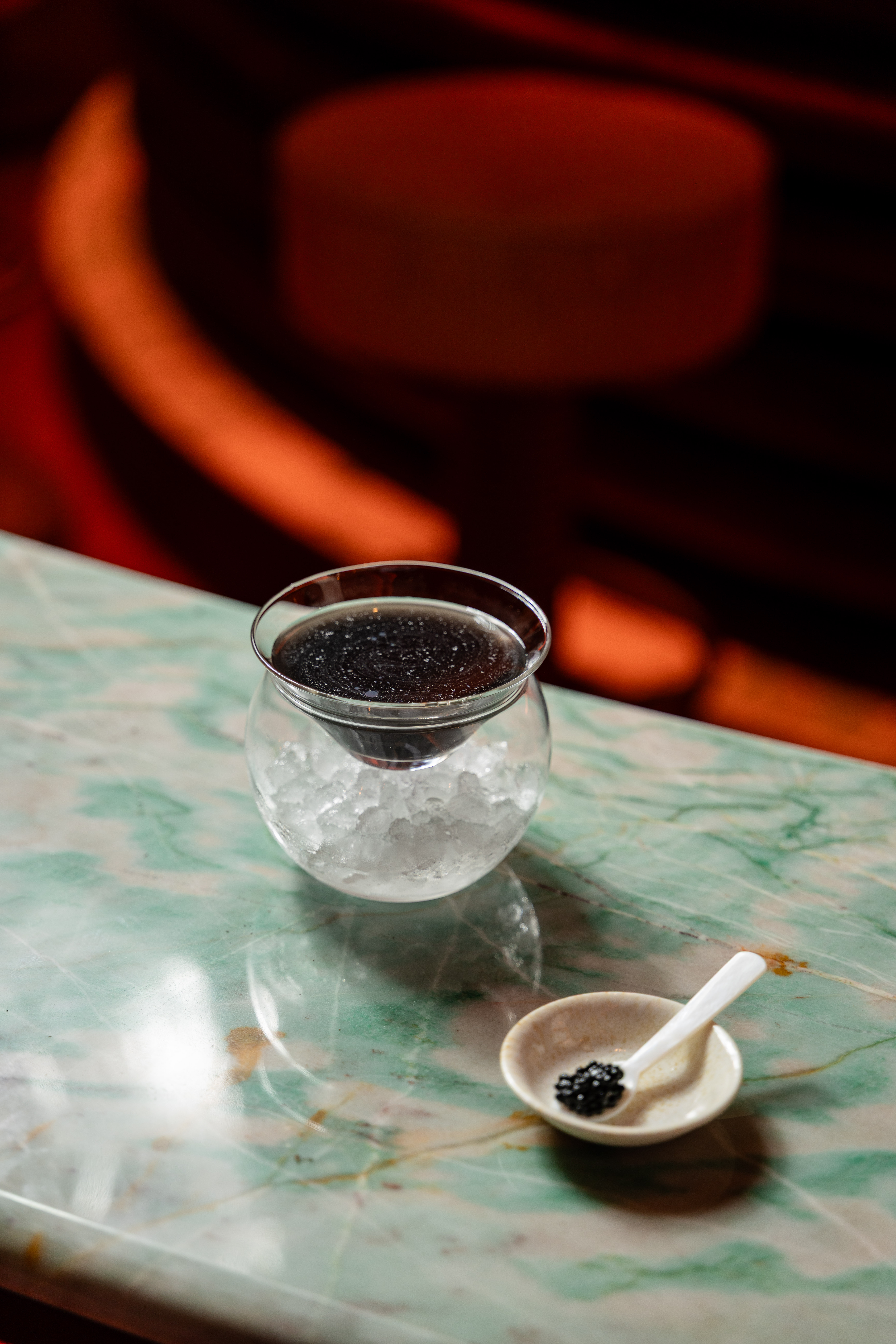 Cocktail servi sur glace pilée dans un verre ballon, accompagné d'une petite cuillère de caviar sur soucoupe, posé sur une table en marbre vert.