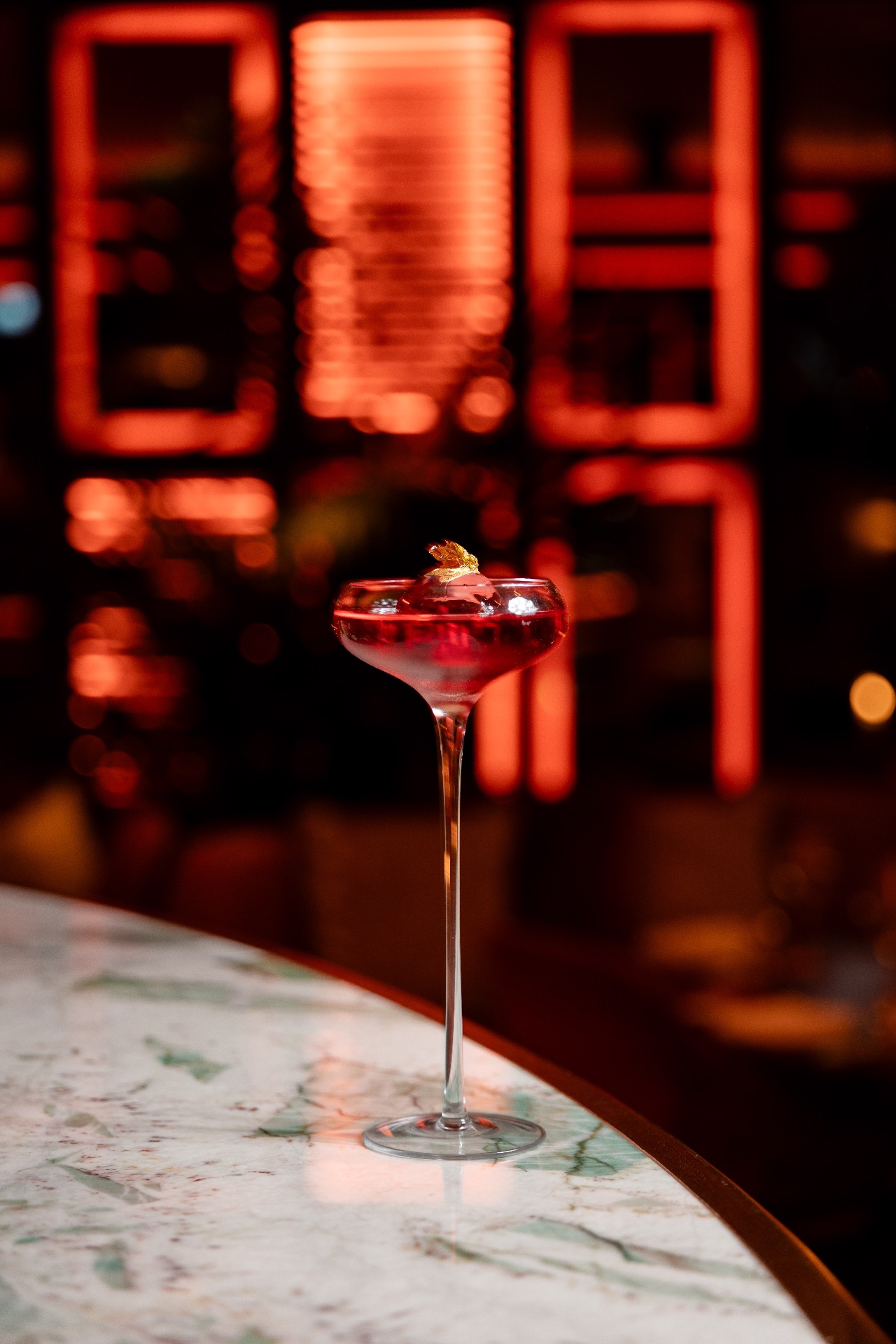 Cocktail rouge servi dans une coupe sur pied, garni d'une décoration dorée, posé sur un comptoir en marbre avec éclairages rouges en arrière-plan.