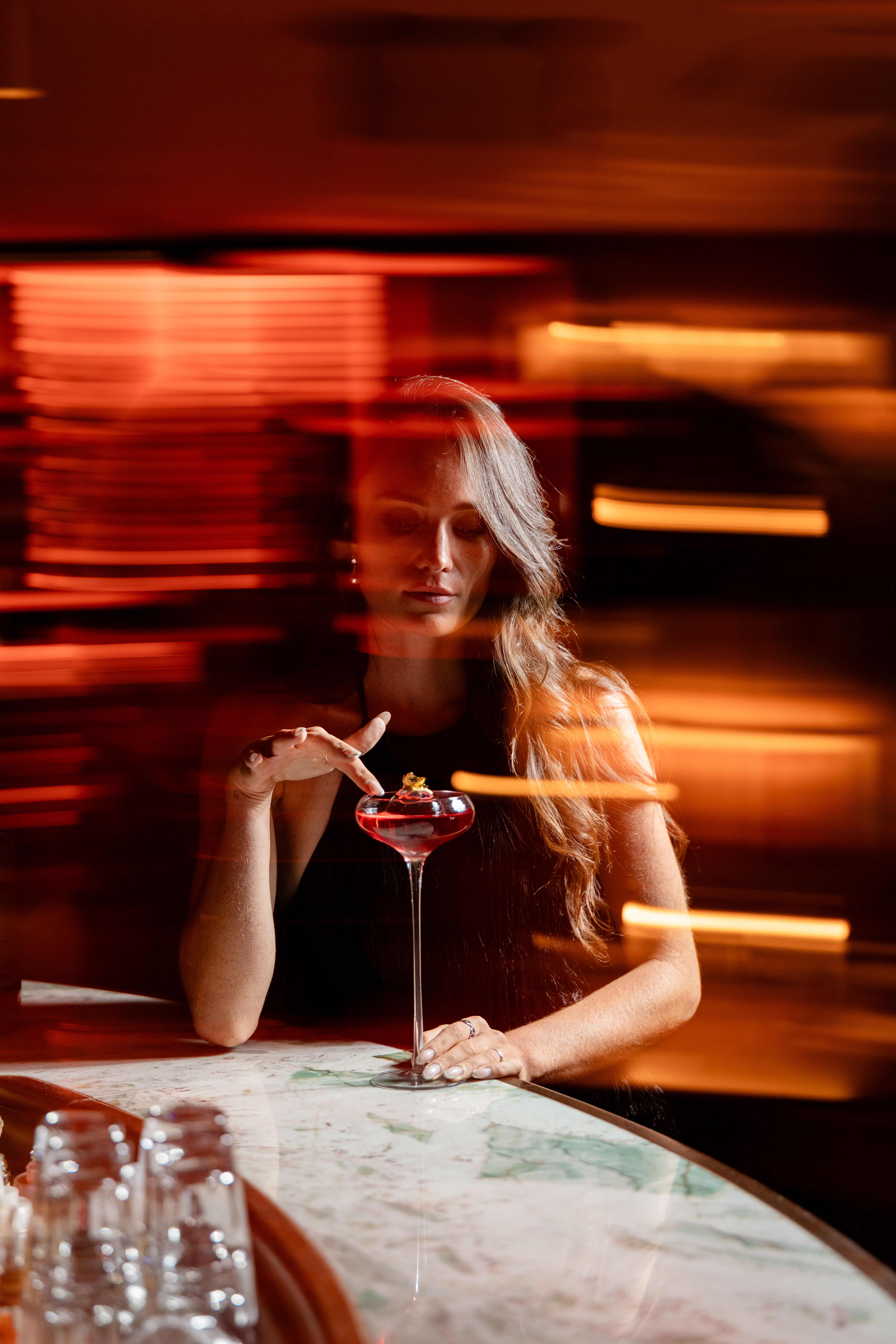 Femme accoudée au bar avec un cocktail rouge devant elle, effet de mouvement flouté et éclairages chaleureux en arrière-plan.