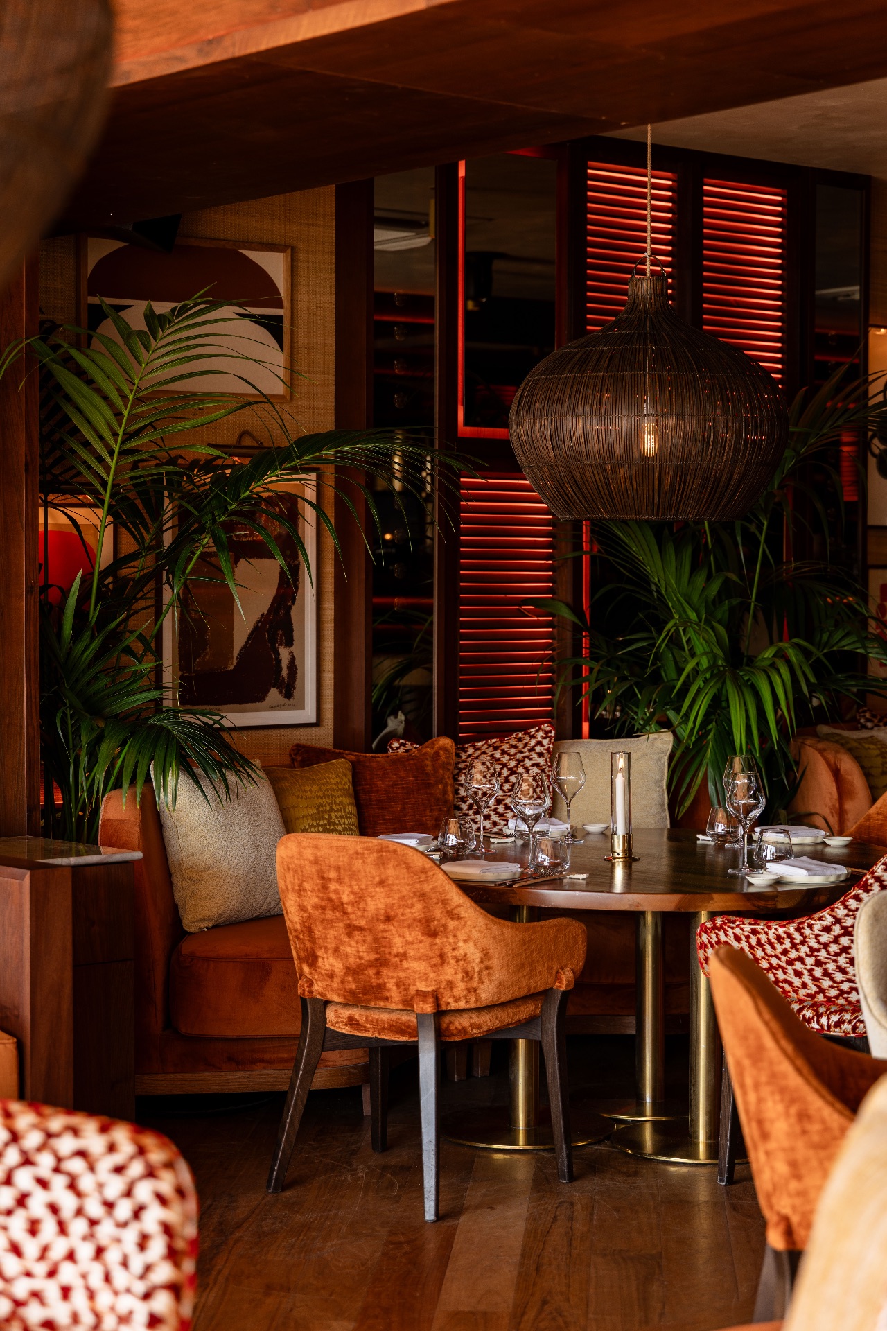 Salle de restaurant intimiste, plantes vertes et suspension en rotin, fauteuils en velours orange autour d'une table dressée, persiennes rétroéclairées en rouge à l'arrière-plan.