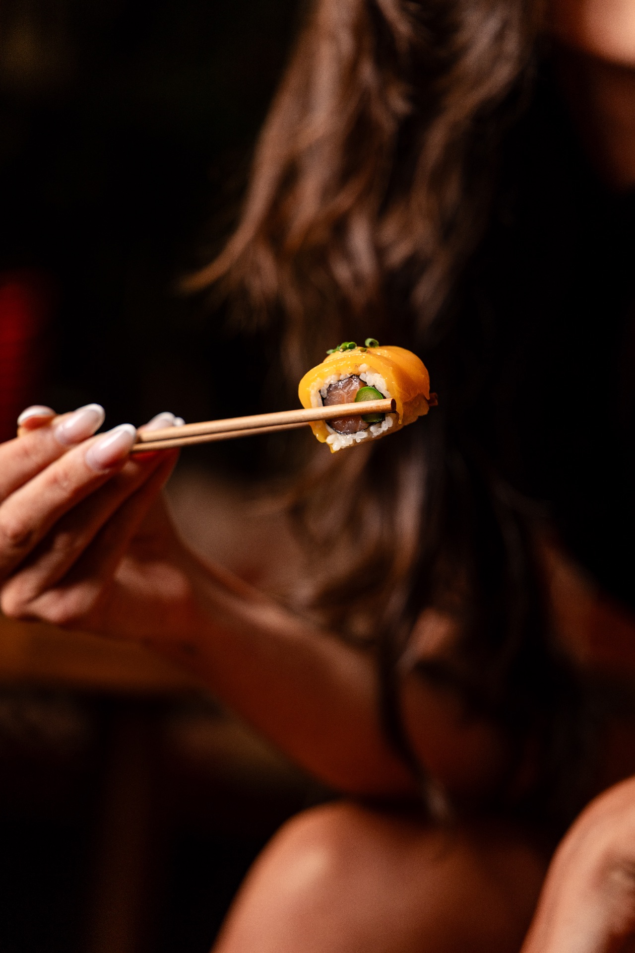 Main tenant avec des baguettes un morceau de sushi maki à la mangue, cheveux au premier plan en arrière-fond flou.