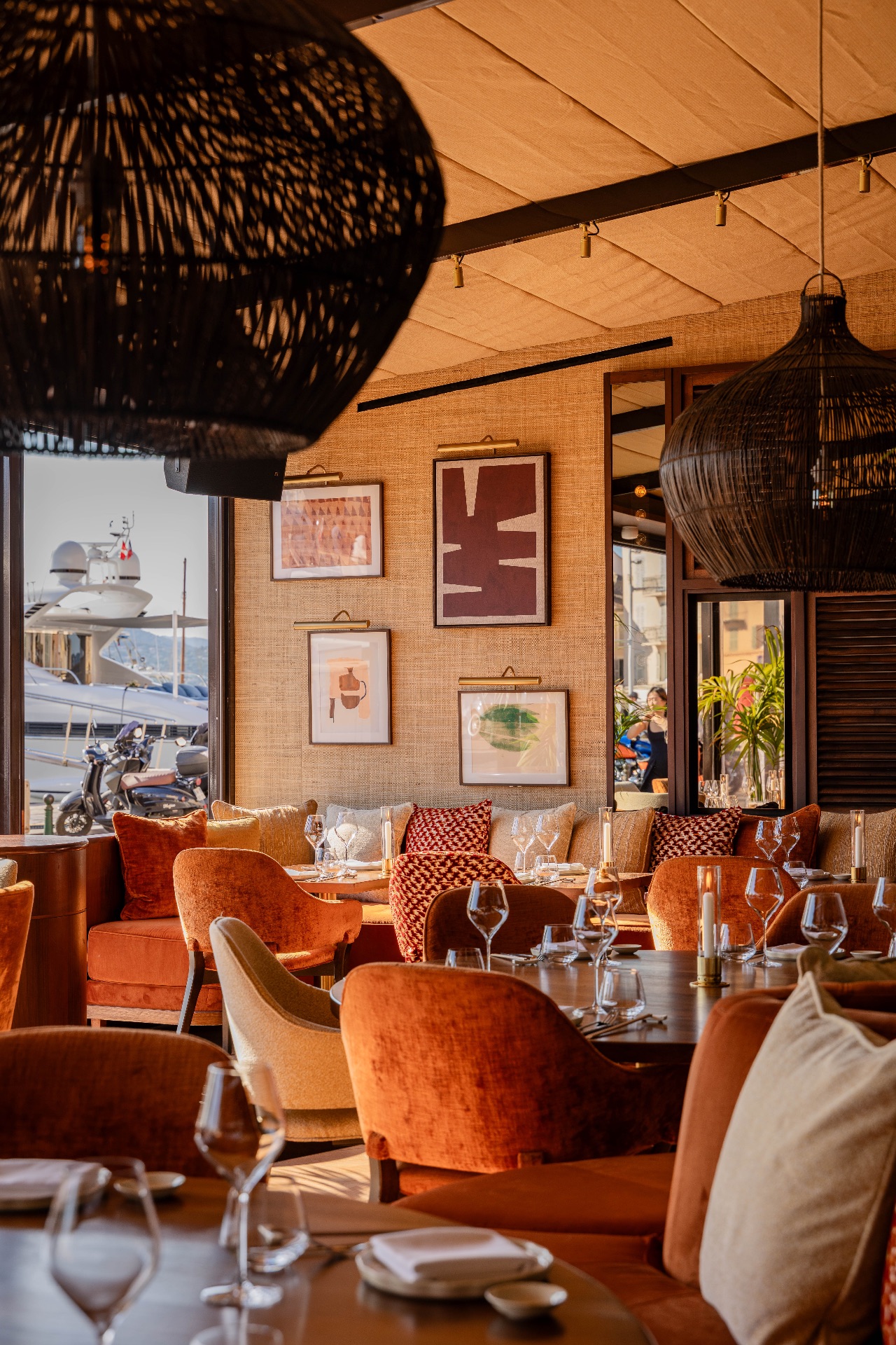 Salle de restaurant baignée de lumière, fauteuils en velours orange autour de tables dressées, suspensions en rotin noir et vue sur le port avec un yacht.