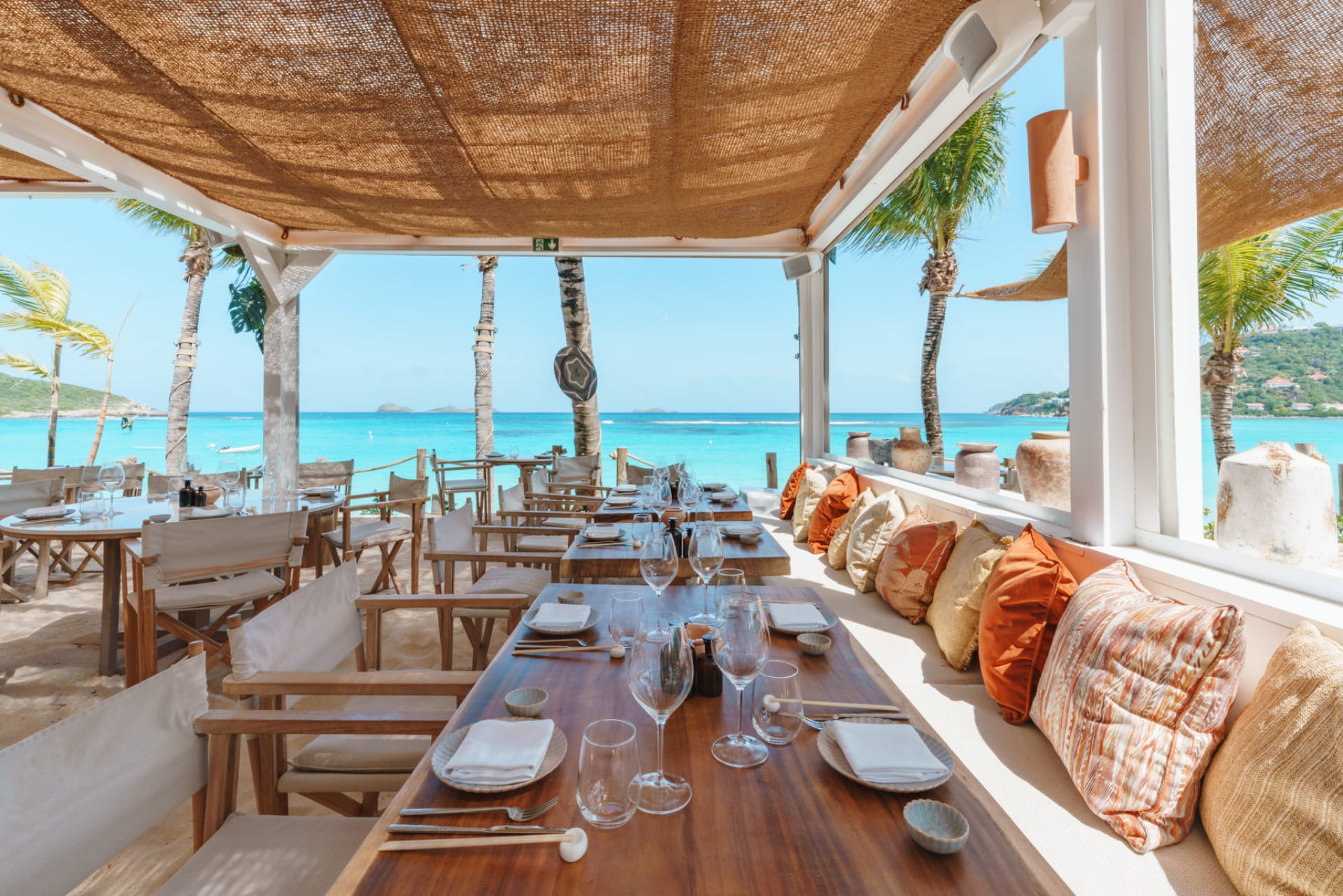 Beach restaurant with set wooden tables, bench seating with colorful cushions, and a view of the turquoise sea lined with palm trees.