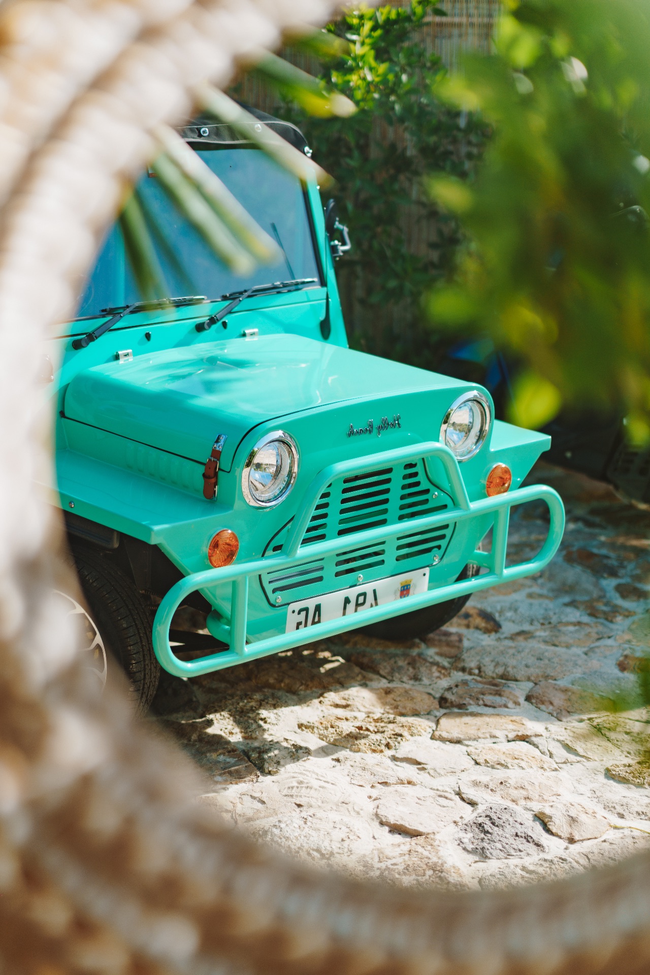 Small vintage turquoise convertible parked on a cobblestone surface, surrounded by foliage.
