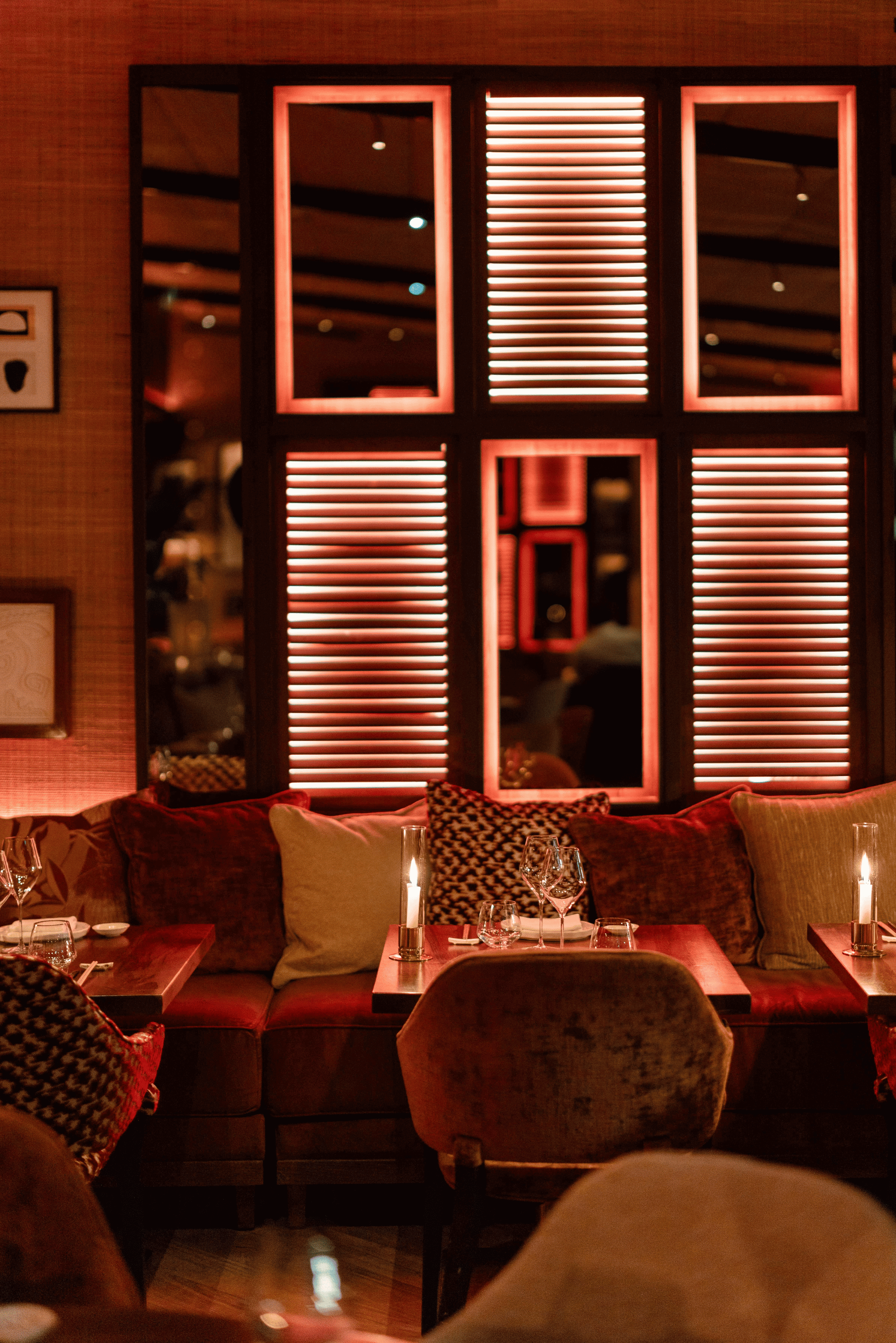 An intimate dining room featuring velvet banquettes with plush cushions, candlelight, and red-backlit shutters.