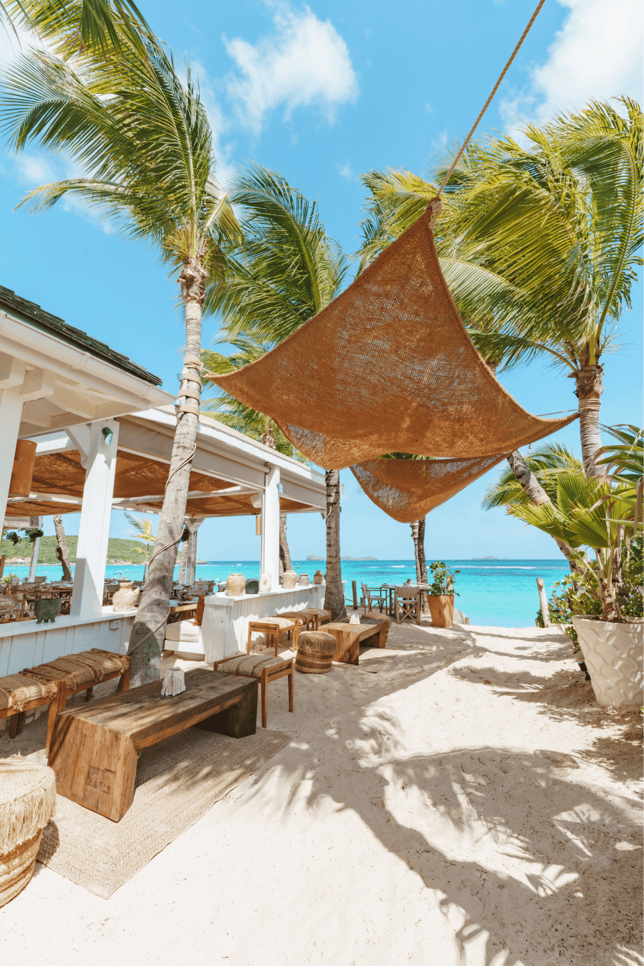 Table dressée en bois sous des palmiers, face à une plage de sable blanc et une mer turquoise avec un bateau au loin.