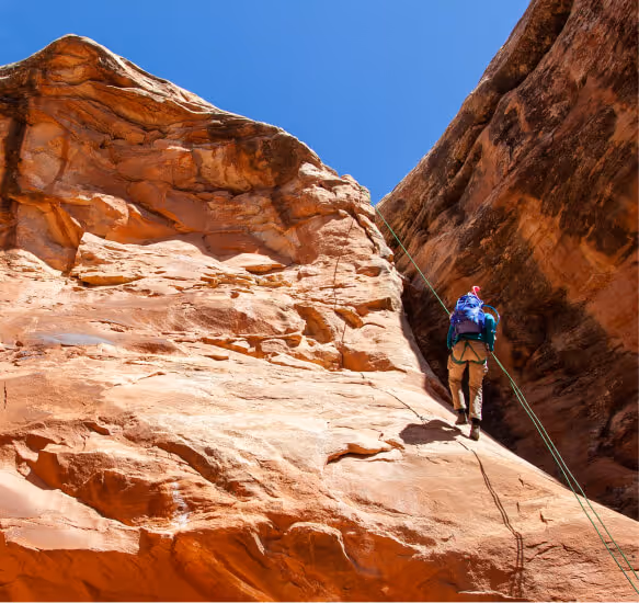 Person rappelling down a steep red rock cliff with clear blue sky background.