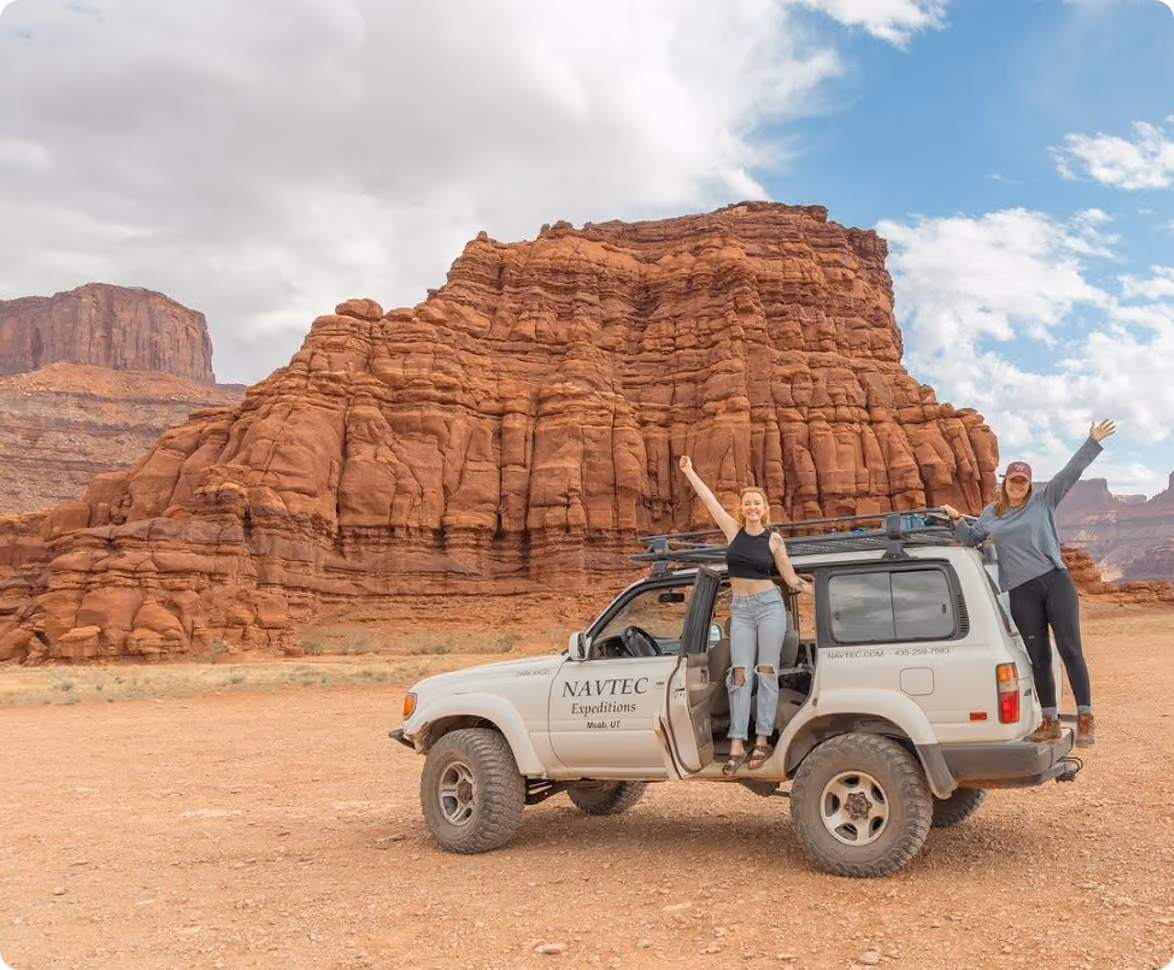 Two women posing on and in a white Navtec Expeditions SUV by red rock formations under a partly cloudy sky.