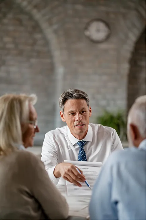 Man in white shirt and tie discussing documents with two older adults in a stone-arched room.