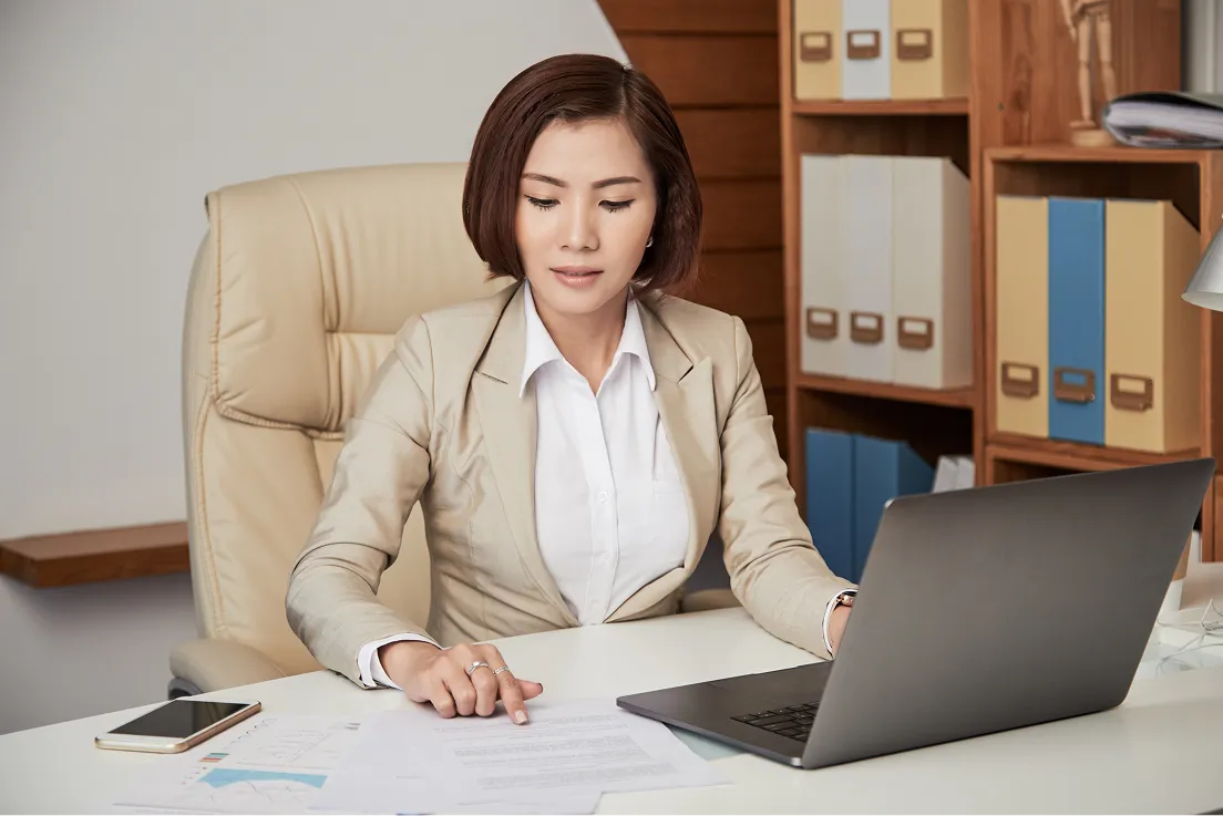 Woman in a beige suit sitting at a desk, reviewing documents with a laptop and phone nearby.
